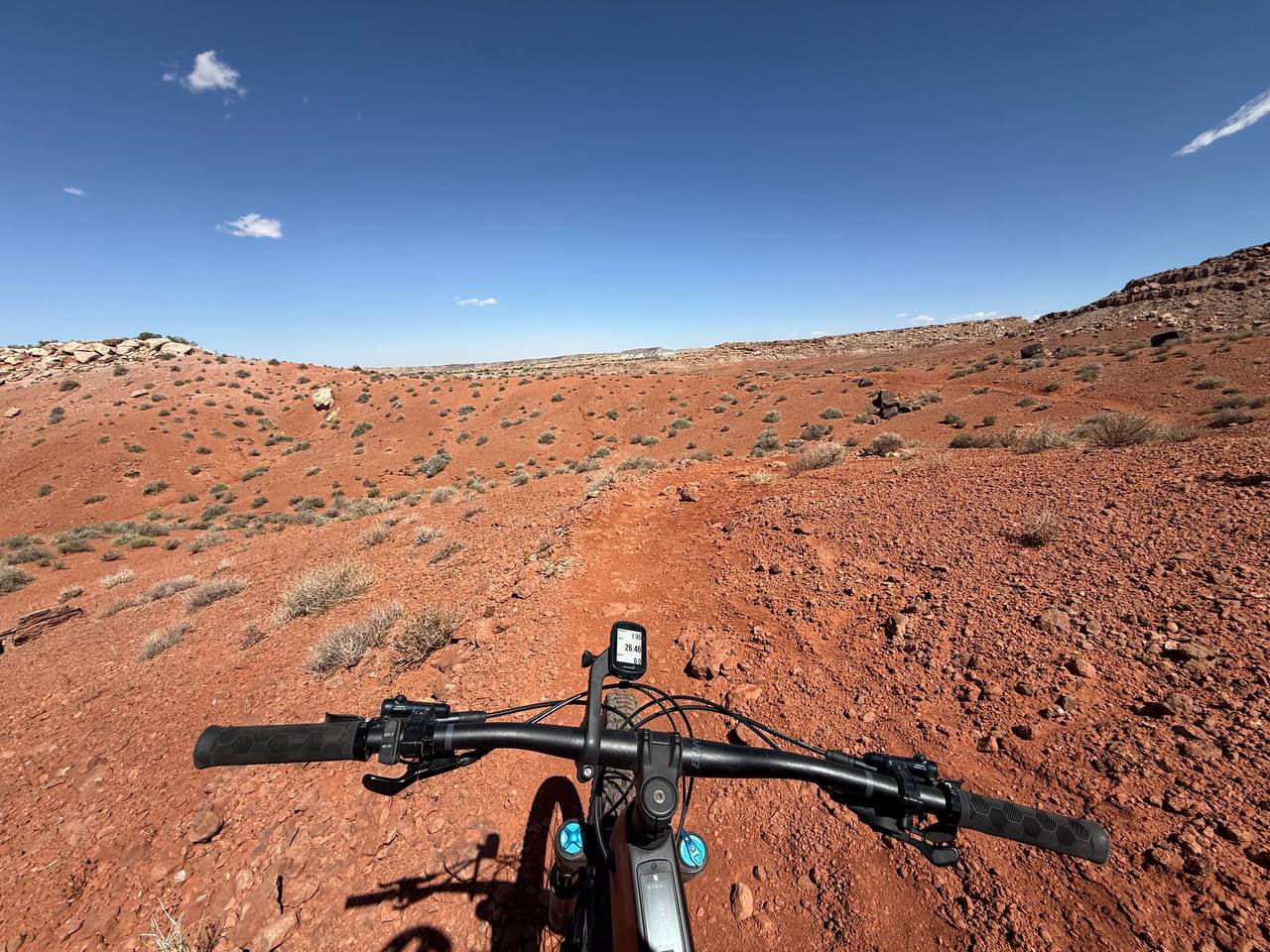 First-person mountain bike handlebar view on a red dirt desert trail under blue sky